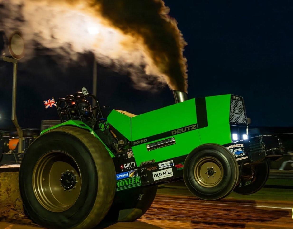 A modified tractor mid-pull under floodlights at night, engine smoking, at an Emley Moor Tractor Pulling event